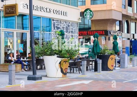 Vue sur la rue avec terrasse du Starbucks Cafe avec vases à fleurs géantes aux visages humains, femme arabe, en premier plan. Banque D'Images