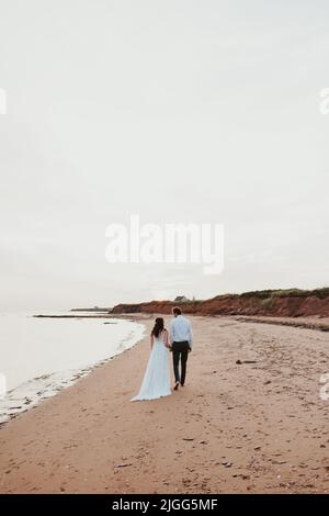 Un cliché vertical d'une mariée et d'un marié marchant sur la plage de sable sous un ciel clair et sans nuages Banque D'Images