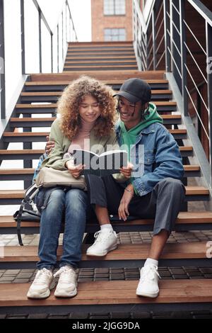 Couple adolescent souriant en vêtements décontractés regardant à travers des notes de conférences dans un livre d'or tout en étant assis sur un long escalier à côté d'un bâtiment moderne Banque D'Images
