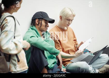 Deux adolescents interculturels regardant à travers des notes de cours dans le copybook tenu par l'un d'eux sur le clavier d'ordinateur portable tout en préparant pour la leçon Banque D'Images