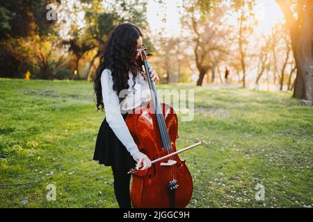 Jeune femme brune debout avec des lunettes jouant du violoncelle au coucher du soleil dans le parc, sur une herbe verte. Banque D'Images