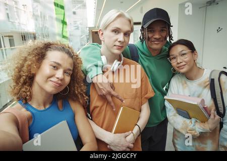 Deux heureux couples adolescents interculturels en vêtements décontractés regardant un appareil photo tout en faisant des selfies dans le couloir de l'université en pause ou après les cours Banque D'Images