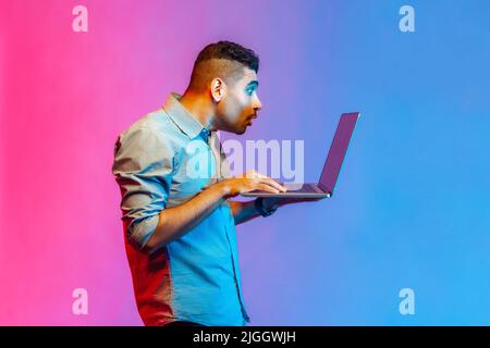 Vue latérale d'un homme en chemise faisant un travail indépendant sur ordinateur portable, en tapant un e-mail ou en surfant sur Internet, regardant surpris à l'écran d'ordinateur portable. Studio d'intérieur isolé sur fond de néon coloré. Banque D'Images
