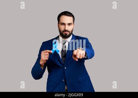 Homme barbu sérieux et confiant tenant un ruban bleu, symbole de la conscience du cancer colorectal, appareil photo pointant du doigt, portant un costume de style officiel. Prise de vue en studio isolée sur fond gris. Banque D'Images