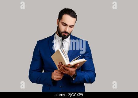 Livre de lecture d'homme barbu concentré, appréciant la lecture, se tenant absorbé par l'intrigue passionnante, vérifier le calendrier dans le bloc-notes, portant le costume de style officiel. Prise de vue en studio isolée sur fond gris Banque D'Images