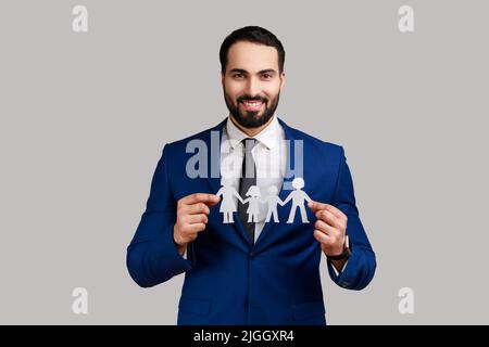 Homme barbu souriant tenant les gens de la chaîne de papier entre les mains, concept de famille heureuse, de parentalité, d'enfance, portant un costume de style officiel. Prise de vue en studio isolée sur fond gris. Banque D'Images