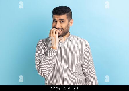 Portrait d'un homme d'affaires barbu stressé qui mord les ongles, nerveux au sujet des problèmes, qui panique et qui a l'air effrayé, portant une chemise rayée. Studio d'intérieur isolé sur fond bleu. Banque D'Images