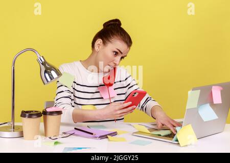 Portrait d'une jeune femme chargée, responsable de bureau, assise sur le lieu de travail, parlant et parlant sur un téléphone fixe, tenant un téléphone portable. Studio d'intérieur tourné isolé sur fond jaune. Banque D'Images