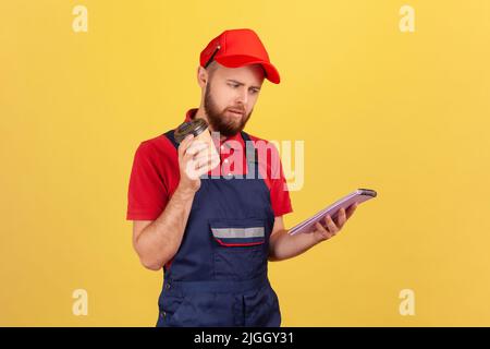 Portrait d'un ouvrier confus portant une combinaison bleue debout avec un carnet en papier et de prendre le café dans les mains, ayant une expression perplexe. Studio d'intérieur isolé sur fond jaune. Banque D'Images