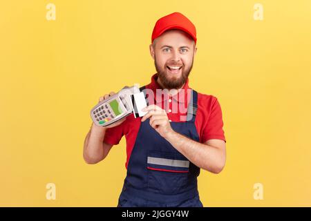 Un homme à barbe enthousiaste qui tient le terminal de pos pour le paiement sans contact et la carte de crédit, payant le service, exprimant le bonheur. Studio d'intérieur isolé sur fond jaune. Banque D'Images