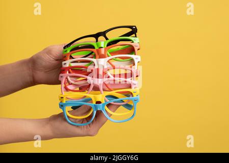 Profil vue latérale gros plan de femme main tenant et montrant beaucoup de différentes couleurs de lunettes. Studio d'intérieur isolé sur fond jaune. Banque D'Images