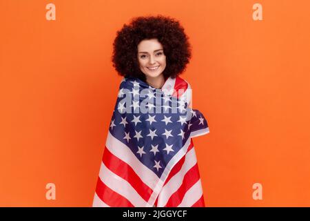 Femme satisfaite de la coiffure afro portant un chandail vert de style décontracté debout étant enveloppé avec le drapeau américain, exprimant des émotions positives. Studio d'intérieur isolé sur fond orange. Banque D'Images