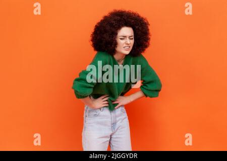 Portrait d'une femme avec une coiffure afro portant un chandail vert de style décontracté souffrant de forte stomachache, frogning, douleur pendant la période. Studio d'intérieur isolé sur fond orange. Banque D'Images