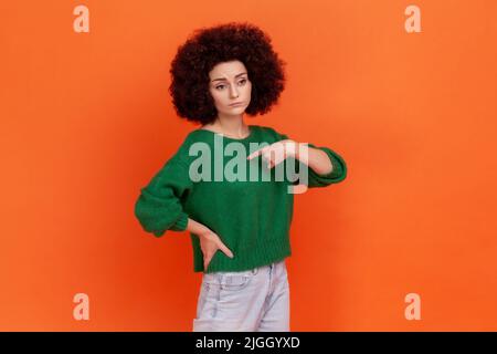 Portrait d'une femme égoïste avec une coiffure afro portant un chandail vert de style décontracté pointant vers elle avec le doigt, ayant l'expression fière. Studio d'intérieur isolé sur fond orange. Banque D'Images