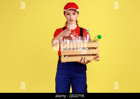 Portrait d'une femme travailleuse sérieuse debout avec une boîte de bouteilles en plastique vides et pointant vers l'appareil photo, vous choisissant, portant une combinaison et un capuchon rouge. Studio d'intérieur isolé sur fond jaune. Banque D'Images