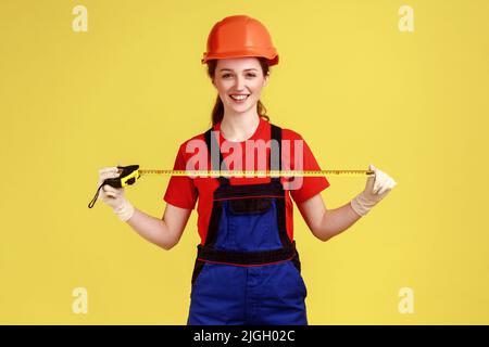 Portrait de femme constructeur tenant la roulette pour mesurer, regardant la caméra avec l'expression du visage heureux, portant des combinaisons et un casque de protection. Studio d'intérieur isolé sur fond jaune. Banque D'Images