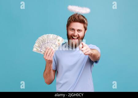 Portrait d'un homme angeliaque barbu satisfait avec Saint nimbus sur la tête montrant le fan de l'argent, tenant des billets de dollars et pointant vers l'appareil photo avec l'index. Studio d'intérieur isolé sur fond bleu Banque D'Images