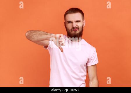 Portrait de l'homme critiquant la mauvaise qualité avec les pouces vers le bas grimace déplaisé, montrant le geste déplaît, exprimant la désapprobation, portant un T-shirt rose. Studio d'intérieur isolé sur fond orange. Banque D'Images