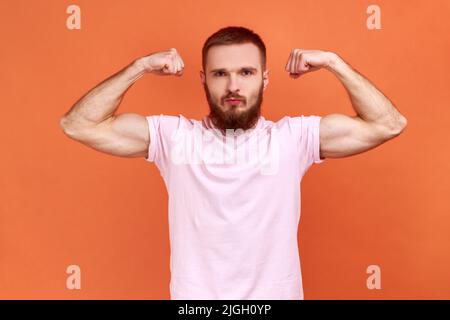 Portrait d'un homme barbu fort démontrant du pouvoir dans ses mains, montrant des biceps, sentant de l'énergie pour gagner le succès, portant un T-shirt rose. Studio d'intérieur isolé sur fond orange. Banque D'Images