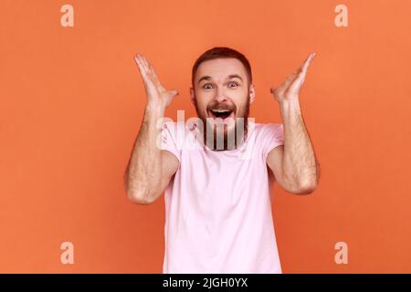 Portrait d'homme barbu debout avec la bouche ouverte dans la surprise, a choqué l'expression, entend des nouvelles incroyables, les mains levées, portant le T-shirt rose. Studio d'intérieur isolé sur fond orange. Banque D'Images