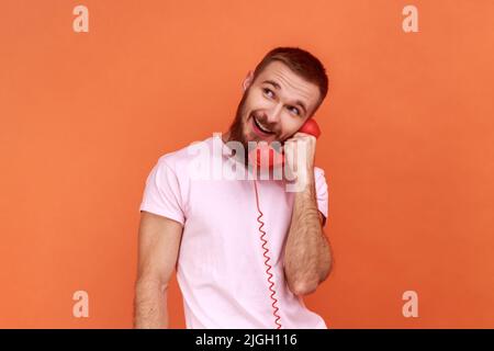 Portrait d'un homme barbu parlant téléphone fixe tenant dans le combiné, regardant loin avec l'expression rêveuse, en portant un T-shirt rose. Studio d'intérieur isolé sur fond orange. Banque D'Images