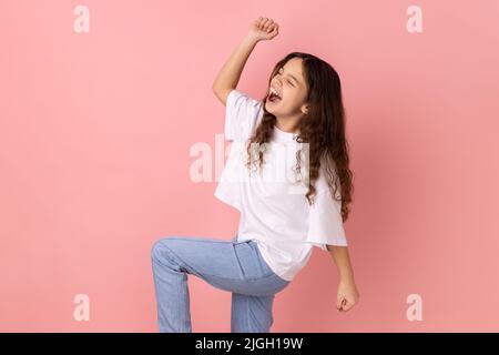 Heureux satisfait petite fille portant T-shirt blanc montrant oui geste et criant célébrant sa victoire, succès, rêves est vrai, euphorie. Studio d'intérieur isolé sur fond rose. Banque D'Images