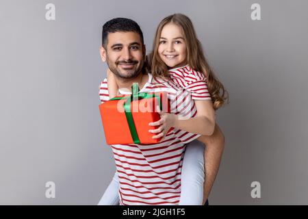 Portrait de père et de fille heureux en T-shirts rayés debout avec boîte de cadeau, enfant préparant le cadeau pour la journée de père, exprimant des émotions positives. Prise de vue en studio isolée sur fond gris. Banque D'Images