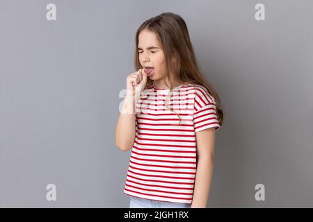 Portrait de malade malsain petite fille à cheveux foncés portant un T-shirt rayé ayant des symptômes de grippe, toux, gardant la main près de la bouche. Prise de vue en studio isolée sur fond gris. Banque D'Images