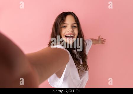 Portrait d'une petite fille portant un T-shirt blanc prenant selfie, regardant l'appareil photo POV et montrant le geste de bienvenue, point de vue de la photo. Studio d'intérieur isolé sur fond rose. Banque D'Images