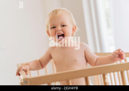 Portrait de drôle de bébé blond de 11 mois debout dans le lit tenant sur des pare-chocs criant avec enthousiasme heureux de voir son père de retour à la maison de travail. Une enfance heureuse et insouciante Banque D'Images