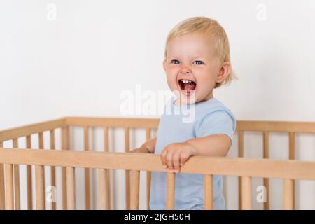 Bébé de dix mois debout dans un berceau avec une expression du visage joyeuse et excitée, en essayant de marcher en tenant sur les butoirs de lit. Une enfance insouciante Banque D'Images