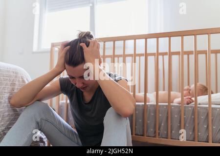 Jeune mère tenant la tête dans les mains avec désespoir, se sentant fatigué et épuisé, pleurant assis à côté de bébé dormant dans le lit, souffrant de dépression postnatale dans le besoin de traitement psychologique Banque D'Images