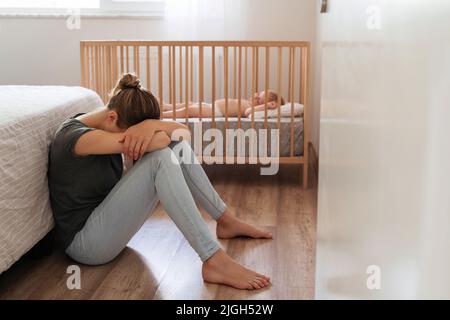 Jeune mère souffrant de dépression postnatale assise sur le sol à côté de bébé qui se couche dans le lit, se reposant la tête sur les genoux, pleurant, se sentant désespérément à la recherche d'un soutien psychologique professionnel Banque D'Images