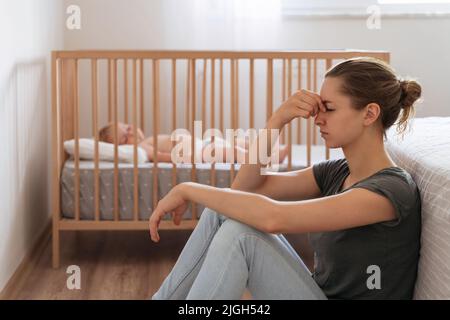 Vue latérale d'une jeune mère frustrée et malheureuse assise sur le sol dans la chambre d'enfant pendant que bébé dormait au lit, essayant de se calmer, souffrant de symptômes de dépression postnatale Banque D'Images