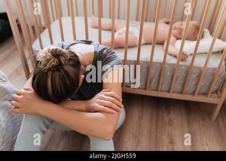 Jeune femme aux difficultés de la maternité assise à côté de bébé berceau avec la tête sur ses genoux pleurant avec l'épuisement et le désespoir, se sentant solitaire. Symptômes de dépression postnatale Banque D'Images