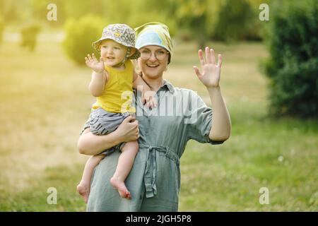 La mère tient un bébé garçon heureux dans ses bras et fait un vagues de sa main. Enfant souriant avec maman qui fait bonjour ou bye geste. Enfant d'un an Banque D'Images