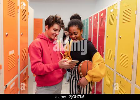 Les jeunes élèves du secondaire se tenant près d'un casier dans le couloir du campus qui parlent et utilisent un téléphone intelligent. Banque D'Images