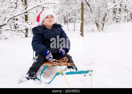 Un beau garçon dans un chapeau de Père Noël est assis sur un traîneau et sourit dans la forêt enneigée d'hiver Banque D'Images