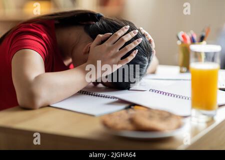 Une jeune fille asiatique triste et fatiguée se couvre des oreilles avec les mains et se trouve sur une table à l'intérieur de la chambre Banque D'Images