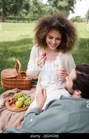 bonne femme maurique tenant la fraise et un verre de vin près d'un petit ami souriant pendant le pique-nique Banque D'Images