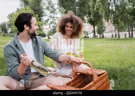 homme gai prenant une bouteille du panier en osier près de la femme maurique tenant la planche à découper avec des fruits pendant le pique-nique Banque D'Images