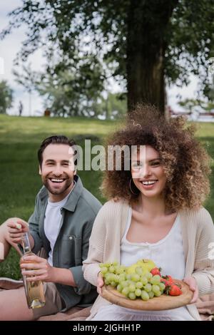 joyeux homme ouvrant une bouteille de vin près d'une petite amie bouclés souriante, assis avec des fruits pendant le pique-nique Banque D'Images