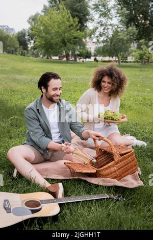 joyeux homme prenant la bouteille du panier en osier près de la petite amie maurique tenant la planche à découper avec des fruits pendant le pique-nique Banque D'Images