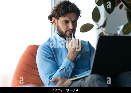 Homme d'affaires d'âge moyen, concentré et réfléchi, utilisant un ordinateur portable, ayant des problèmes avec le travail dans un bureau moderne Banque D'Images