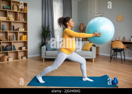 Souriant jolie jeune femme enceinte noire faisant des exercices avec le ballon de fitness sur le tapis, profiter de la maternité Banque D'Images