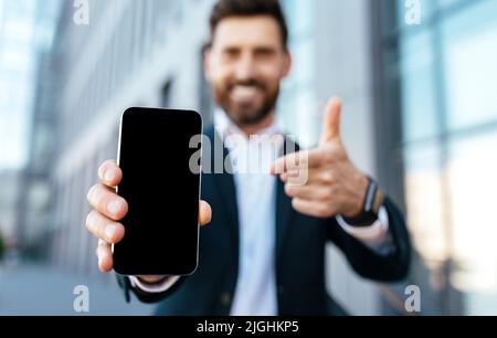 Un jeune homme d'affaires européen souriant et barbu en costume se réjouit de la réussite et pointe le doigt sur un smartphone Banque D'Images