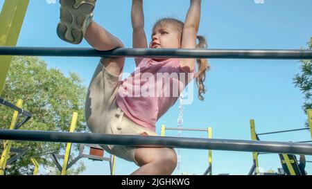 Une petite fille monte l'échelle de gymnastique sur un terrain de sport ouvert à l'extérieur. Une petite fille mignonne à ramper sur une échelle de sport verticale dans le parc de la ville le jour du soleil. Banque D'Images