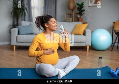 Maternité et équilibre aquatique. Une jeune femme afro-américaine souriante est assise sur un tapis et de l'eau potable Banque D'Images