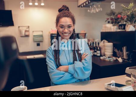 Bonne belle jeune femme élégante avec des tresses debout au comptoir dans le café et souriant. Banque D'Images