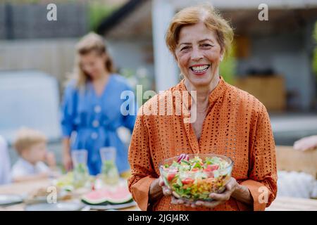 Bonne femme senior qui sert de la salade lors de la fête de jardin de plusieurs générations en été. Banque D'Images